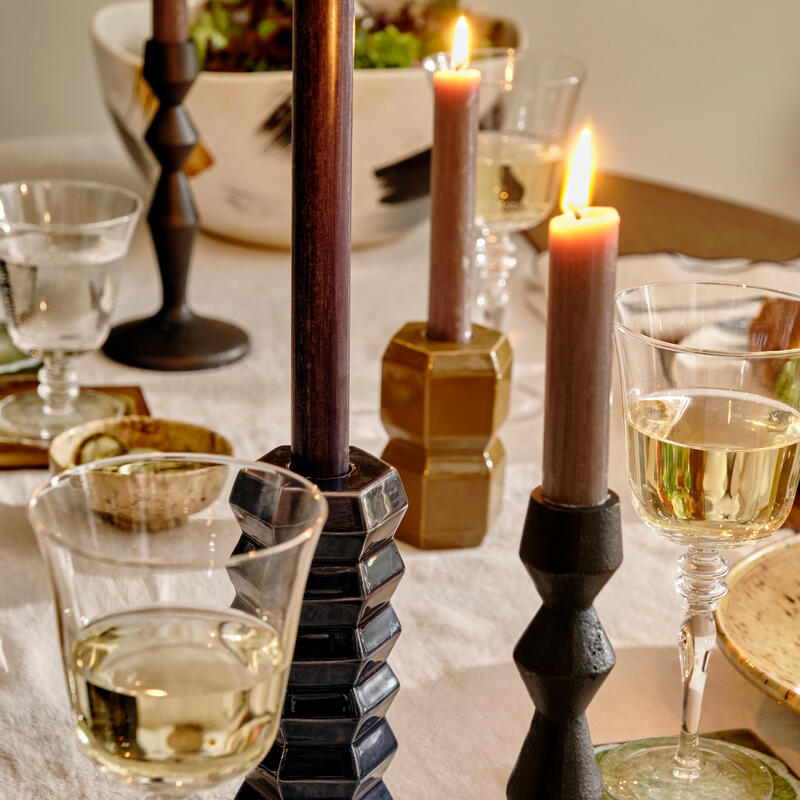 Candles are burning in geometric holders atop a white tablecloth, surrounded by wine glasses and a bowl of salad, creating a cozy dining setting.