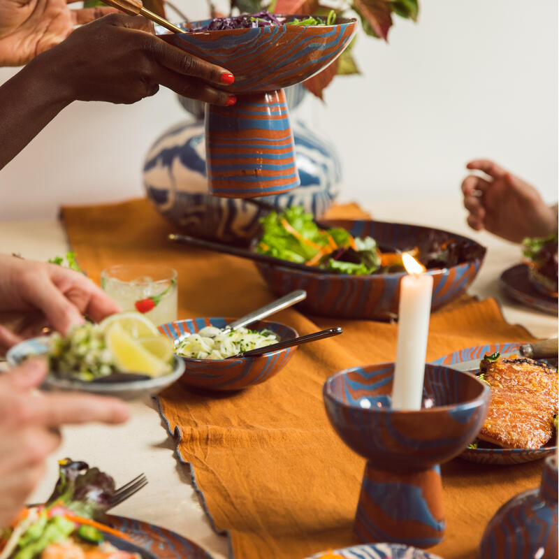 Hands serve food from colorful bowls, including salad and grilled meat, around a table set with candles and drinks. The table is covered with an orange cloth, and a decorative vase is visible in the background.