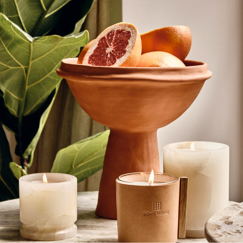 Wax candles burn on a marble table near a terracotta bowl filled with grapefruits and lush green leaves; "SOHO HOUSE" is engraved on a glass candle holder with a leather cover.