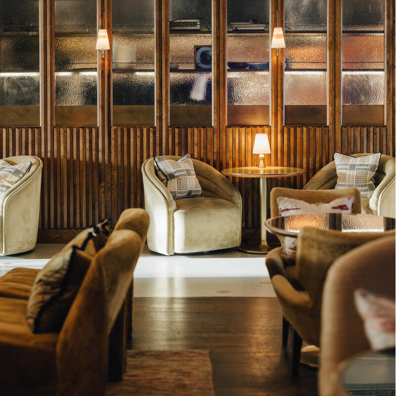 Plush chairs arranged around small tables hold patterned pillows in a cozy lounge. Dim lamps illuminate a textured wood and glass wall, casting warm light in the elegant setting.