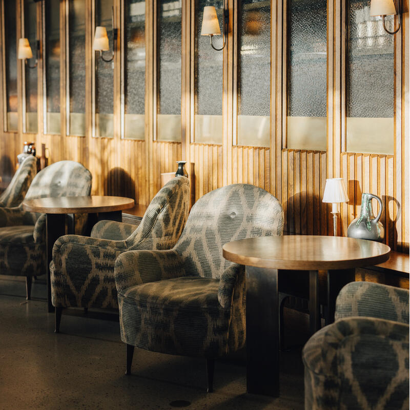 Armchairs with patterned upholstery and round wooden tables are aligned against a textured glass-paneled wall with wooden framing. The space is softly lit by wall-mounted lamps.