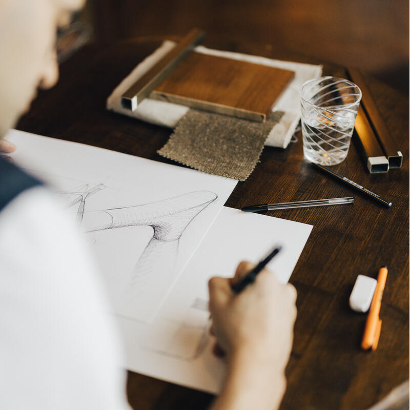 A person sketches a high-heeled shoe design on white paper at a wooden table with drawing tools and a glass of water.