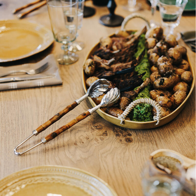 Oval serving tray containing roasted potatoes, lamb chops, and greenery, accompanied by tongs, placed on a wooden table set with glassware, plates, napkins, and utensils in a dining setting.