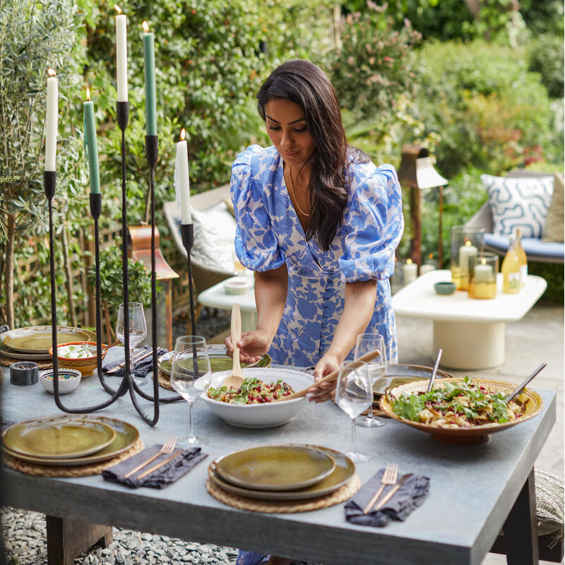 A woman in a blue floral dress serves salad at a candlelit outdoor table, set with plates, glasses, and utensils, surrounded by lush greenery and garden furniture.