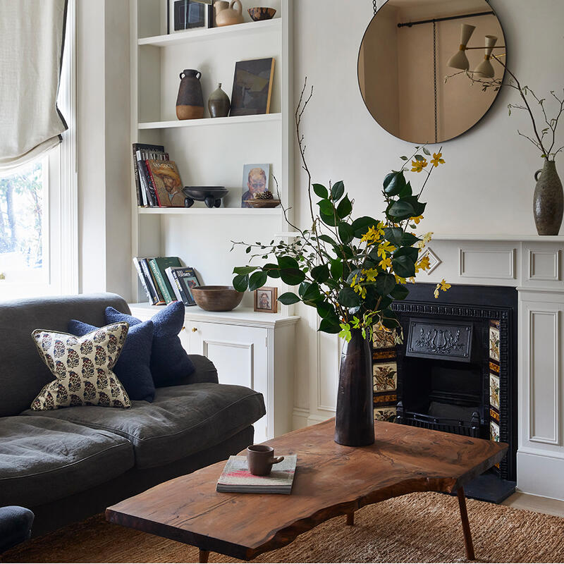 Wooden table holds vase with yellow flowers, books, cup; gray sofa with patterned cushions nearby; wall shelves with decor and books; round mirror above black fireplace; modern chandelier overhead.