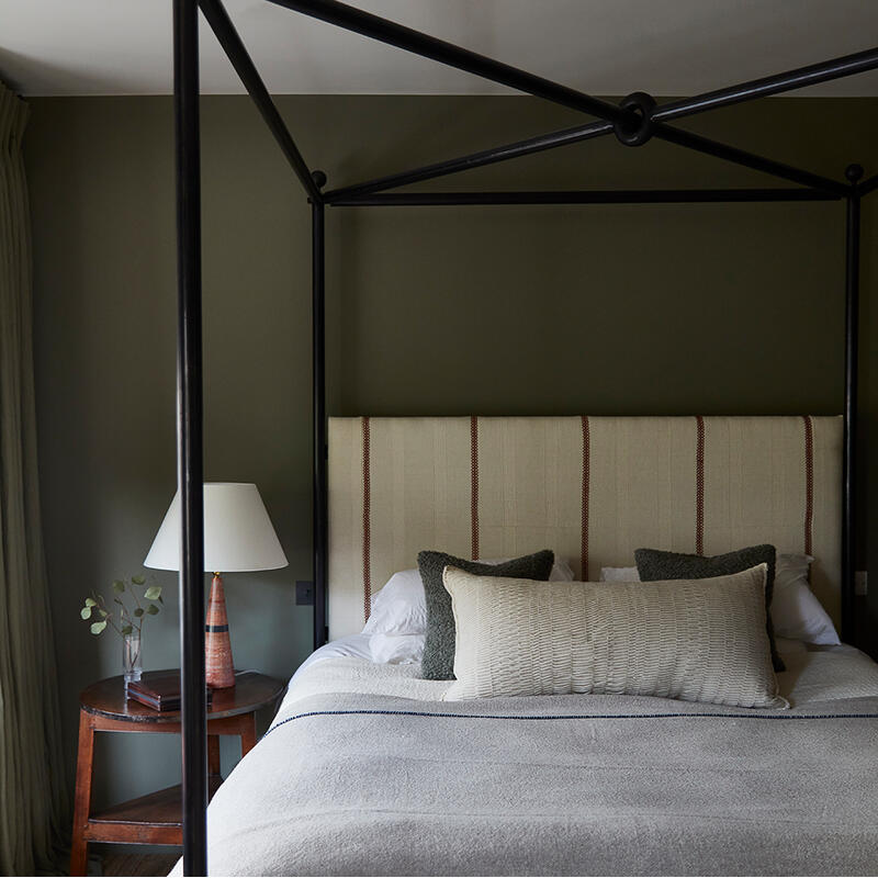 A canopy bed with pillows and a throw blanket stands in a minimalist bedroom, accompanied by a round wooden side table holding a lamp and a glass vase with foliage.