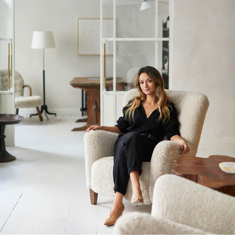 A woman sits on a cream-colored armchair, dressed in black clothing with beige heels, in a minimalist room featuring modern fixtures, large mirrors or glass windows, and additional lounge chairs.