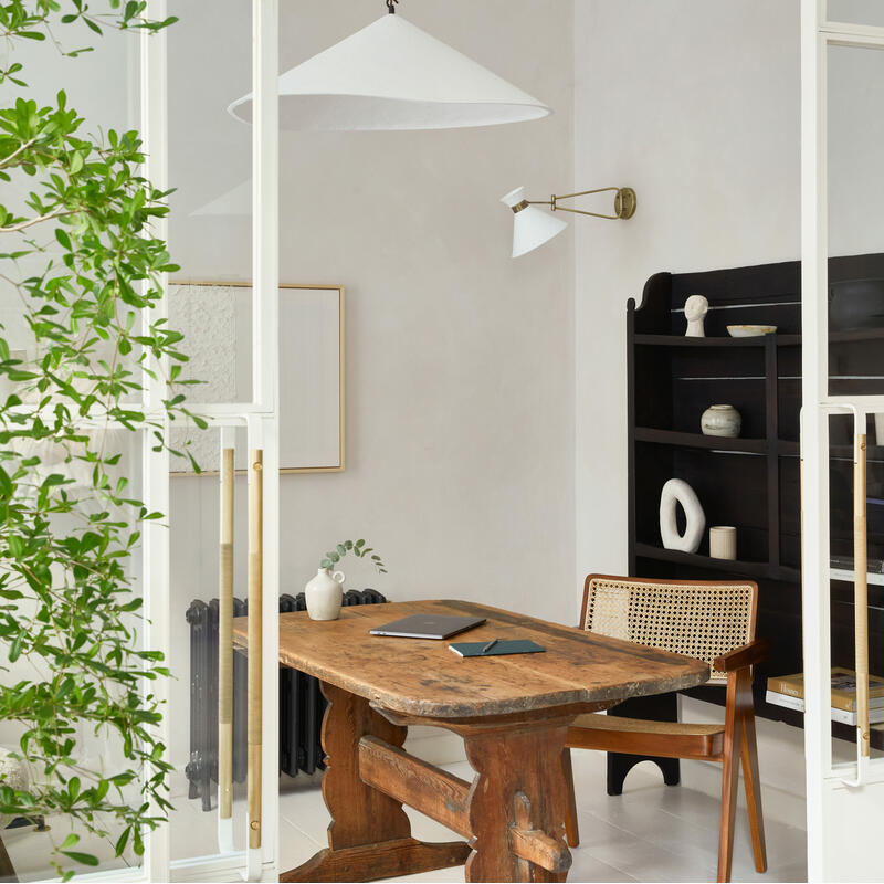 A rustic wooden desk sits with a chair against a light-colored wall adorned with modern art and a wall lamp, near a black bookshelf in a minimalist, plant-adorned room.