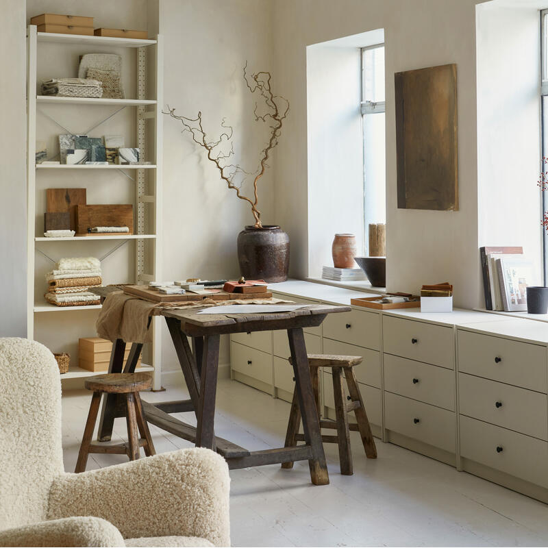 Rustic wooden table and stools in a cozy, well-lit room with neutral colors, surrounded by shelves with books and textiles, drawers, and decorative pottery. A large ceiling lamp and wall art enhance the space.