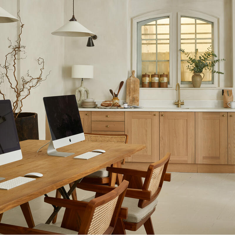 Two desktop computers on a wooden table with chairs in a bright, minimalist kitchen featuring wooden cabinets, rounded window, hanging light fixtures, and countertop items like jars, cutting boards, and plants.