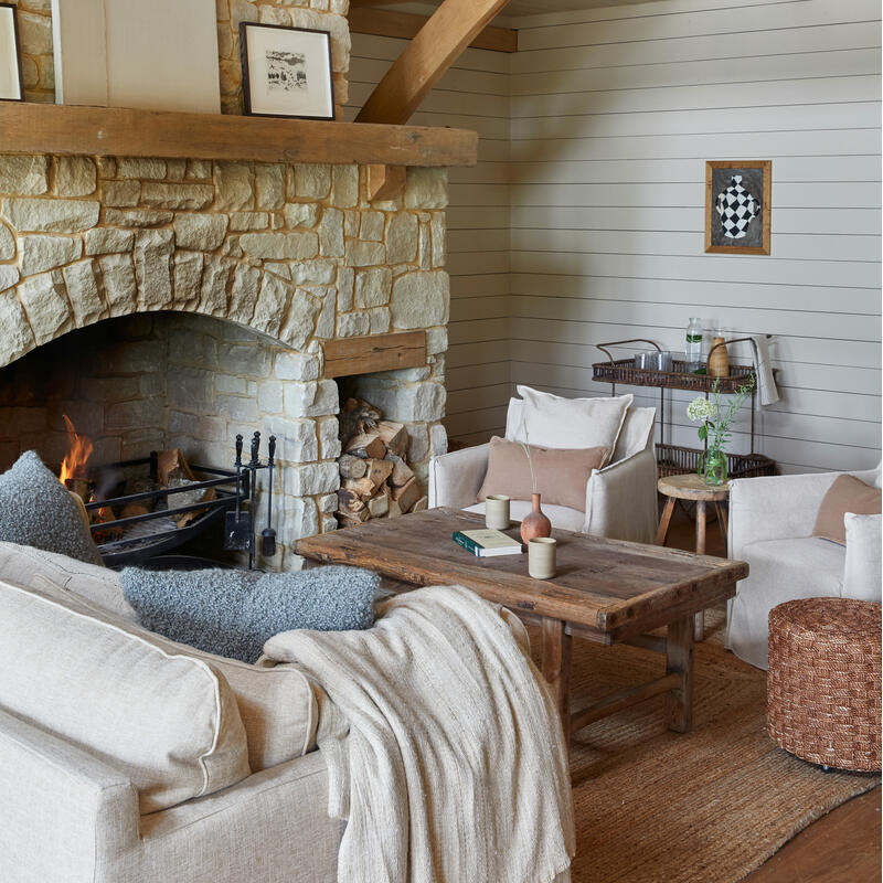 Stone fireplace with a burning fire, in a cozy living room featuring beige sofas, a wooden coffee table, a wicker hanging light, and wooden beams. Artwork and log storage add rustic charm.