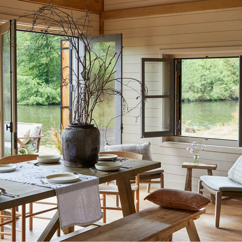 Wooden dining table set with plates, featuring a large vase with twigs, situated in a rustic cabin with open windows and doors, offering a view of a serene river and lush greenery.