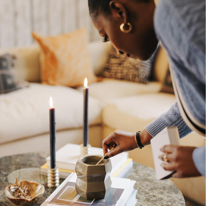 A person lights two black candles on a marble table cluttered with books, a ceramic cup, and a small decorative bowl, in a cozy living room with a beige couch and pillows in the background.