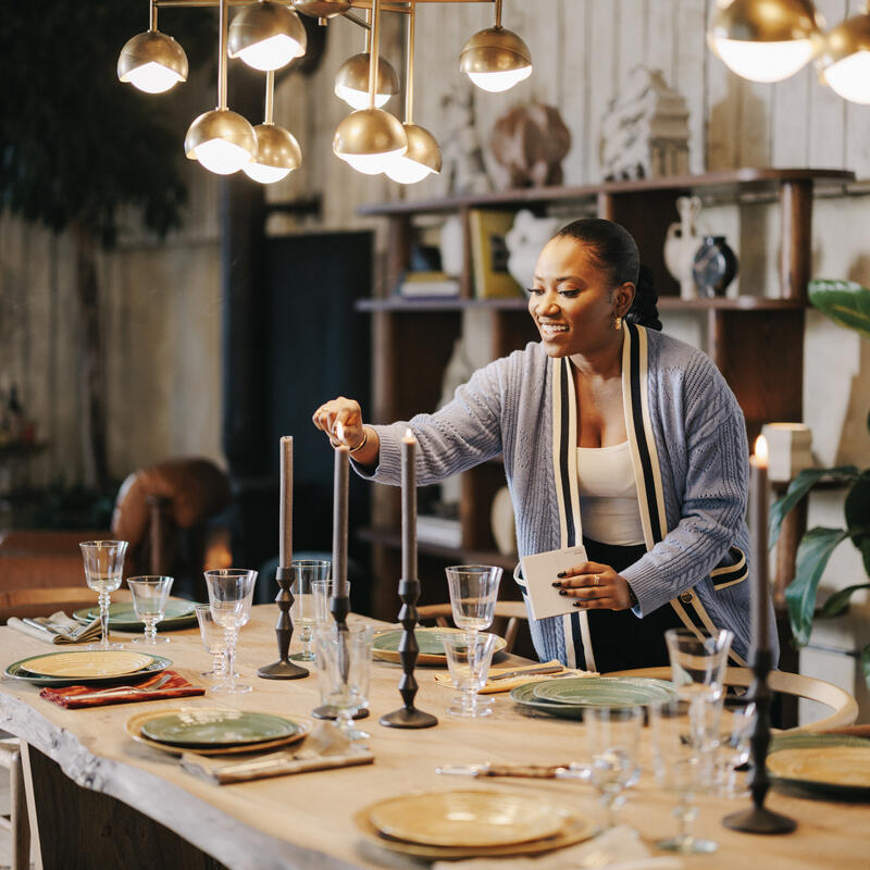 A woman lights candles on a wooden dining table set with plates and glasses, under a chandelier, in a warmly decorated room with bookshelves and plants.