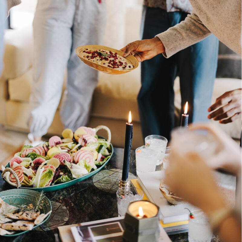 People enjoying food and drinks in a cozy indoor setting, with a focus on a person holding a plate of food near a table adorned with a large platter of salad, candles, and drinks.