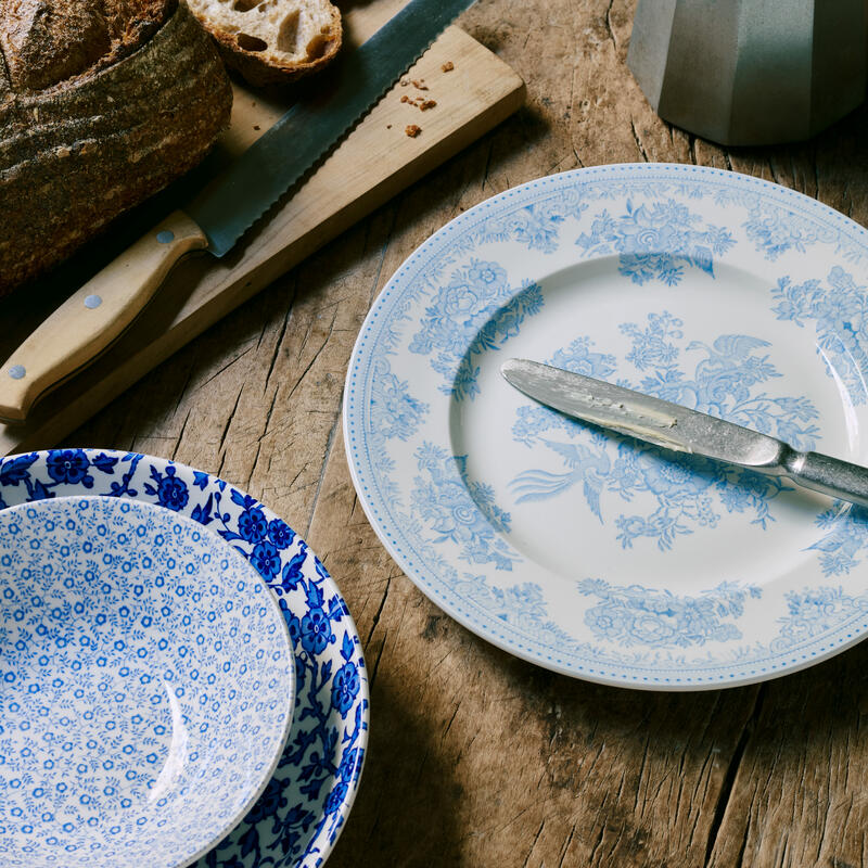 A serrated knife rests on a wooden cutting board with sliced bread near two blue floral-patterned dishes, all placed on a rustic wooden table next to a metal stovetop coffee pot.