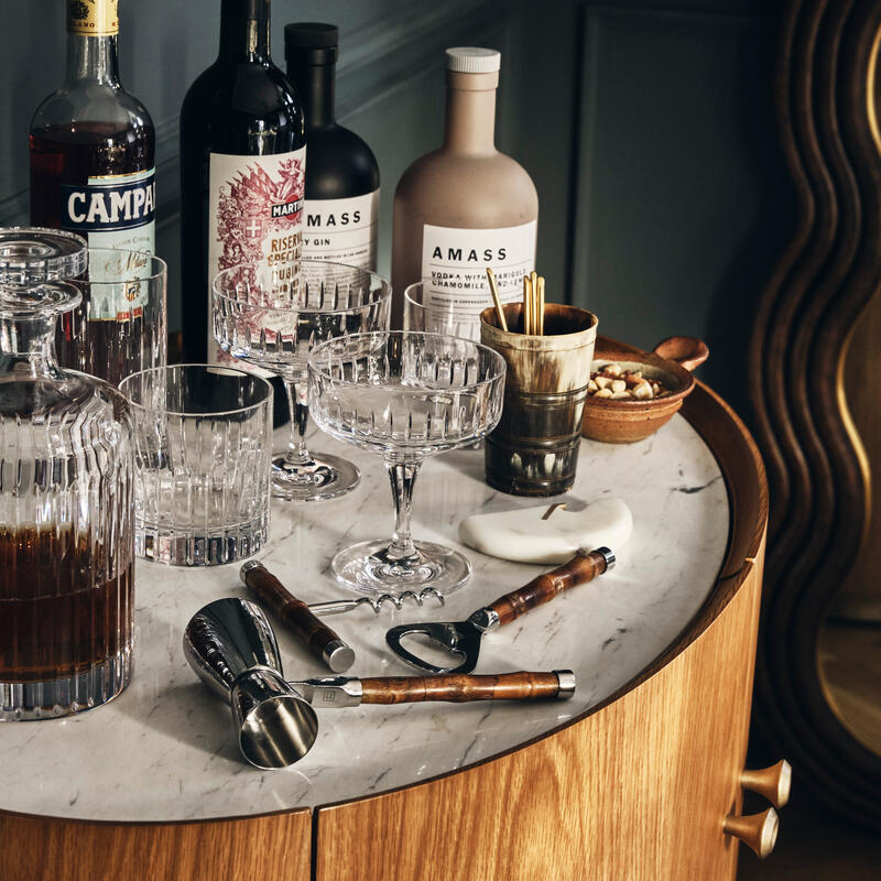 A marble-topped bar cart holds various liquor bottles, crystal glasses, a decanter, bar tools, and a small bowl of nuts, set against a dark wall with a wavy mirror.