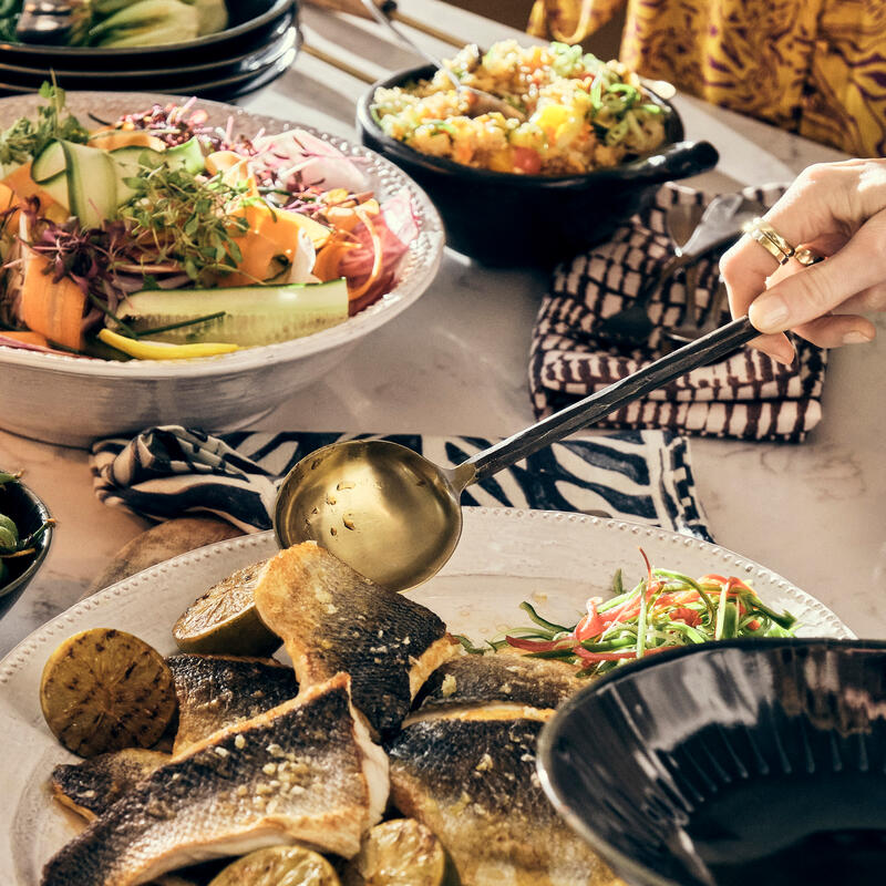 Several dishes are arranged on a table, with a hand ladling food. Grilled fish with lime slices and a colorful salad sit in the foreground, accompanied by a drink.