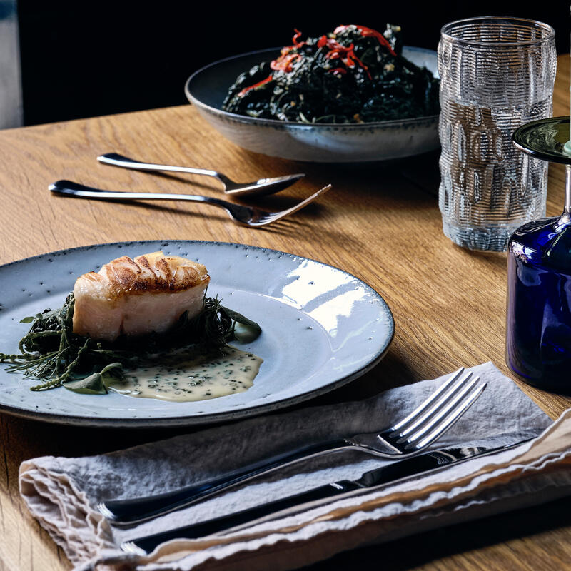 A seared fish fillet atop greens in sauce sits on a ceramic plate on a wooden table, accompanied by utensils, glassware, and a candle.