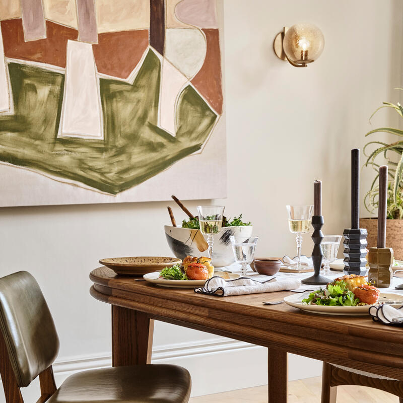 A wooden dining table set with various dishes, glasses, and candles is surrounded by a dark rug and modern chair; abstract wall art and a spherical wall lamp decorate the background.