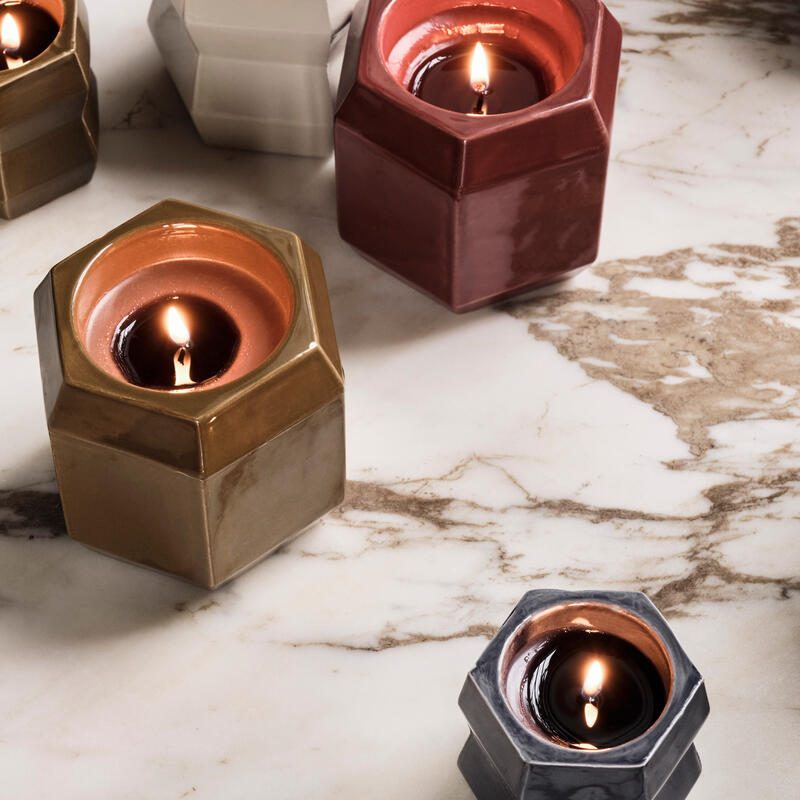 Hexagonal candles flicker on a marbled surface, each housed in colored, glossy holders: white, mustard, maroon, and charcoal grey. The tranquil setting emphasizes a sense of warmth and elegance.