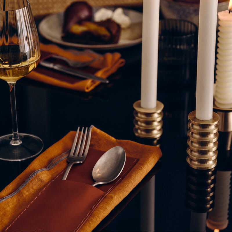 Two lit white candles on brass holders sit on a dark table, beside a glass of wine, a plate with food, and a fork and spoon on a mustard napkin.