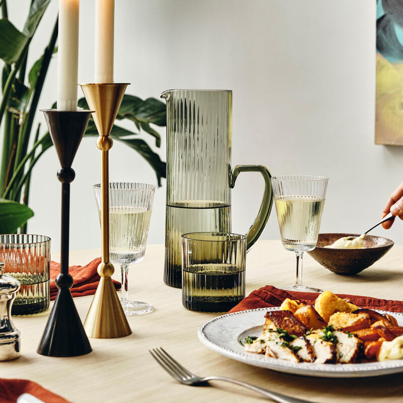 Candles on holders illuminate a dining table set with glassware, a water pitcher, silverware, and plates of food, while houseplants and a wall painting create a cozy, stylish atmosphere.