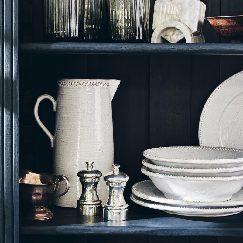 Ceramic pitcher, metallic pepper grinders, and a stack of white dishes sit on dark wooden shelves, surrounded by assorted glassware and decorative items, creating a neatly organized, rustic kitchen display.