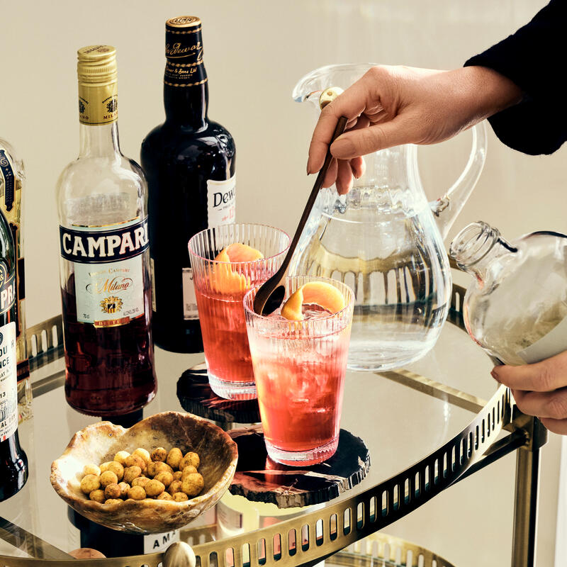 A hand stirs a spoon in a pink cocktail glass garnished with orange slices on a mirrored trolley with liquor bottles, a water pitcher, and a bowl of nuts.