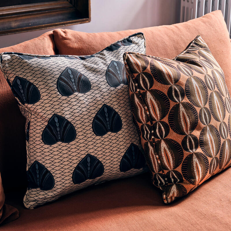 Pillows with intricate patterns rest on a beige fabric sofa in a cozy, likely indoor, setting. The left pillow features heart-shaped designs; the right pillow showcases circular motifs.