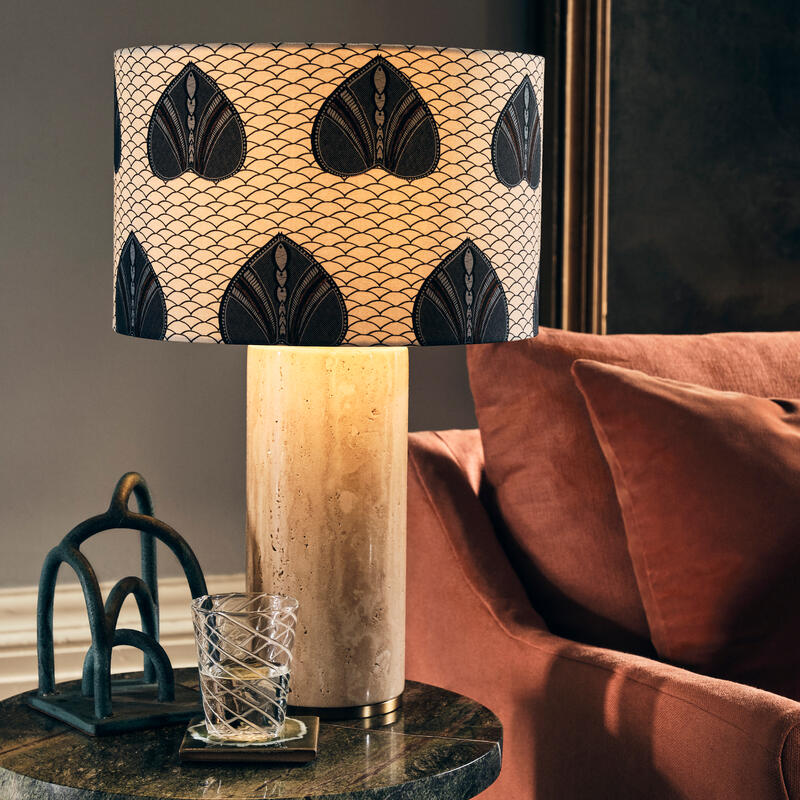 A stone lamp with a patterned shade rests on a side table with a glass and a decorative metal piece, beside a brown sofa in a cozy, dimly lit room.