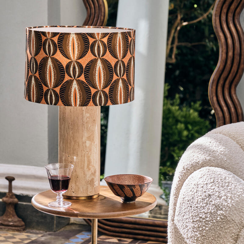 A glass of red wine and a decorative bowl rest beside a patterned lampshade on a wooden side table, set in a tiled room with a wavy-framed mirror and white columns.