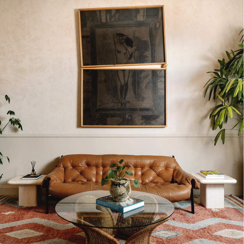 A tan leather couch sits against a wall with a large, split painting above it, flanked by potted plants. A glass coffee table with books and a small plant is in front, all on a patterned rug.