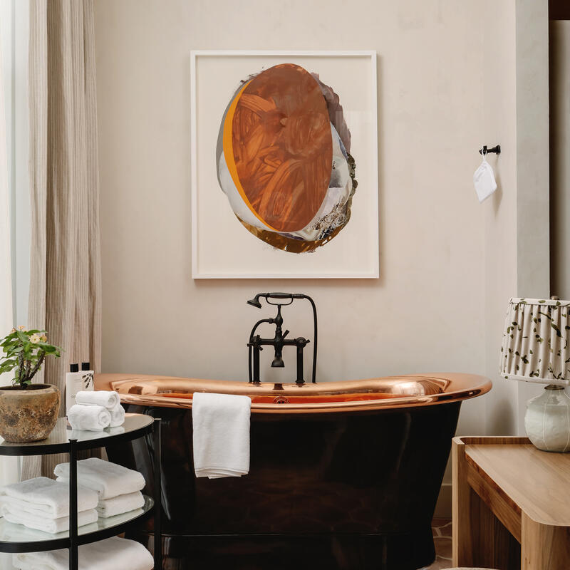 A black bathtub with copper interiors, adorned with white towels, stands against a beige wall featuring abstract art. Nearby, a shelving unit with white towels and a plant, and a wooden bench with a patterned stool sit on a tiled floor.