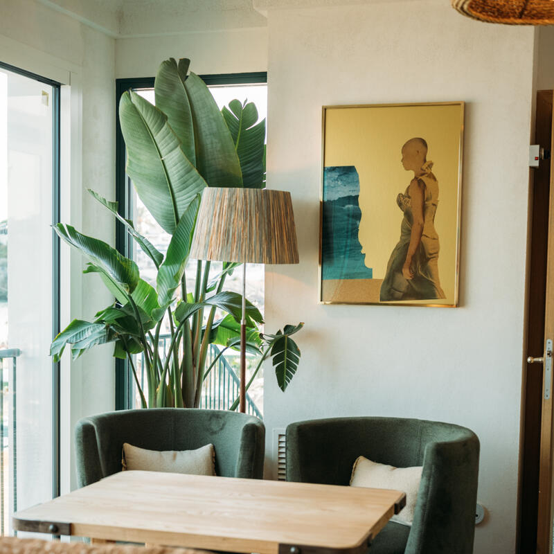 A cozy corner features two green cushioned chairs around a wooden table, large leafy plants, a lamp, and a framed artwork on a white wall, in a sunlit room.