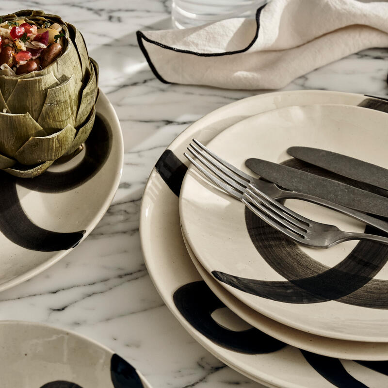 Dinner plates with bold black brushstroke designs hold stacked cutlery, set on a marble countertop. A stuffed artichoke rests on one plate, with a folded napkin and glass of water nearby.