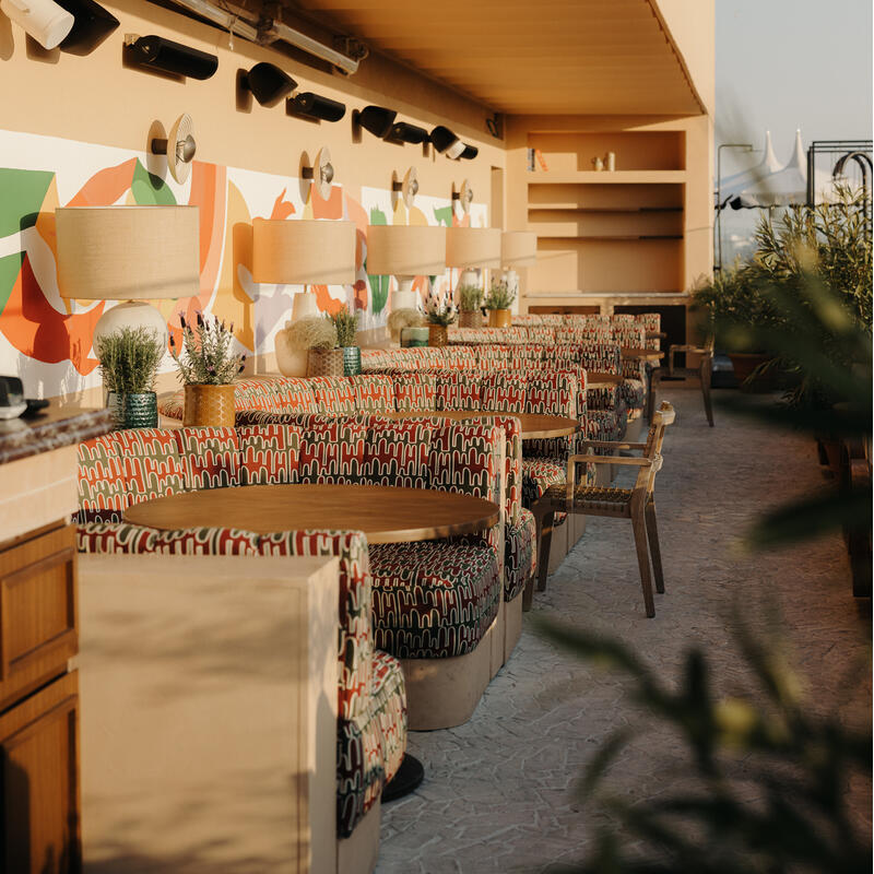 Booths with patterned cushions and wooden tables line an open-air restaurant with warm lighting, abstract wall art, and plants in decorative pots along the wall.