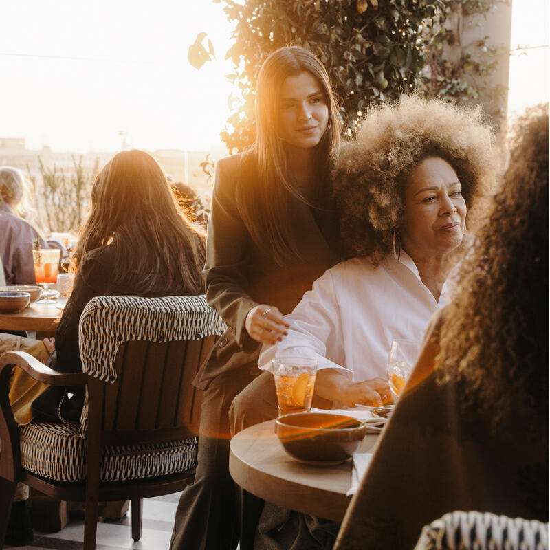 A woman stands beside another seated woman holding a drink, surrounded by several people dining at an outdoor restaurant with lush greenery and sunlight filtering through.