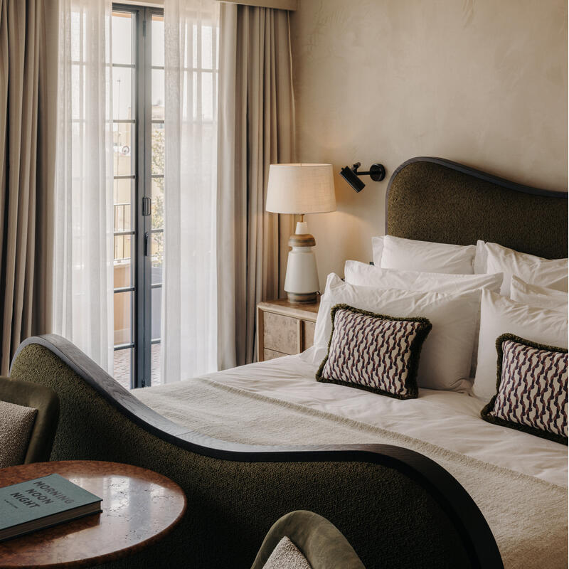 A stylish bedroom showcases a neatly made bed with green accents, bedside table and lamp, book on a round table, and soft natural light filtering through sheer curtains on a window.