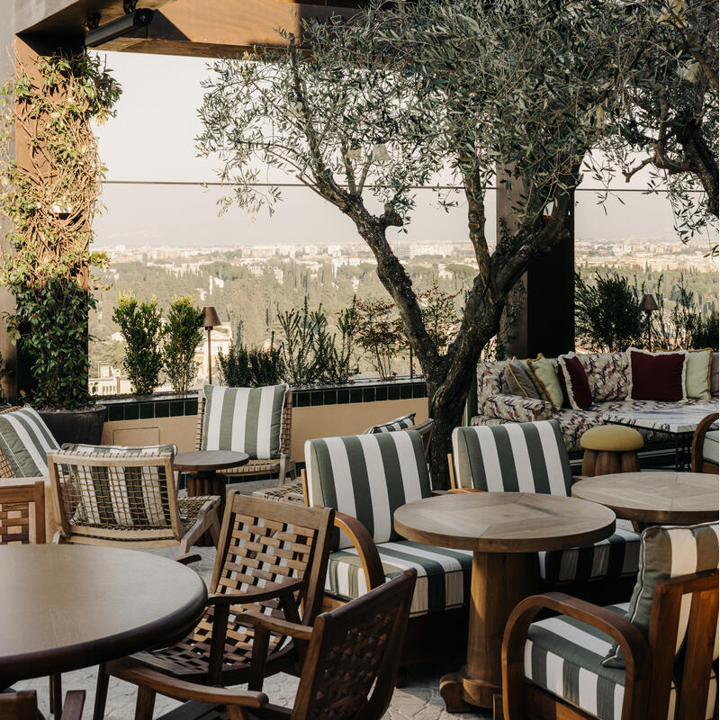 Wooden chairs and tables are arranged in an outdoor lounge area with striped cushioned seating, surrounded by greenery and overlooking a distant cityscape under a wooden pergola.