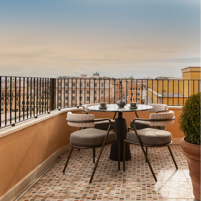 A small round table with chairs and tea settings sits on a tiled balcony overlooking a cityscape with buildings under a clear blue sky.