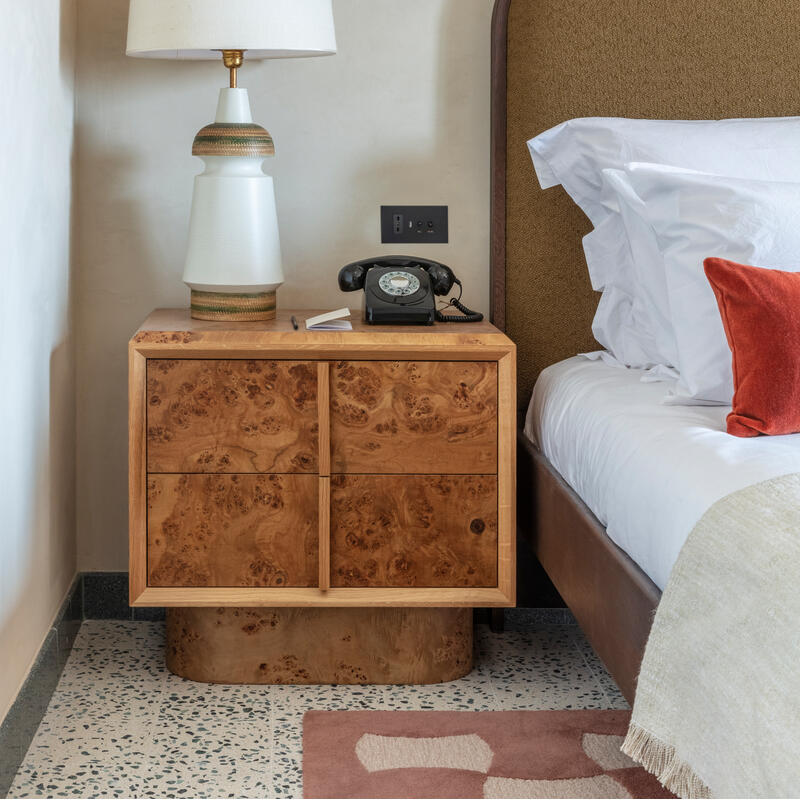 A bedside table holds a ceramic lamp, rotary telephone, and notepad, positioned next to a bed with white linens and red cushion. A rustic wall sconce is mounted above.