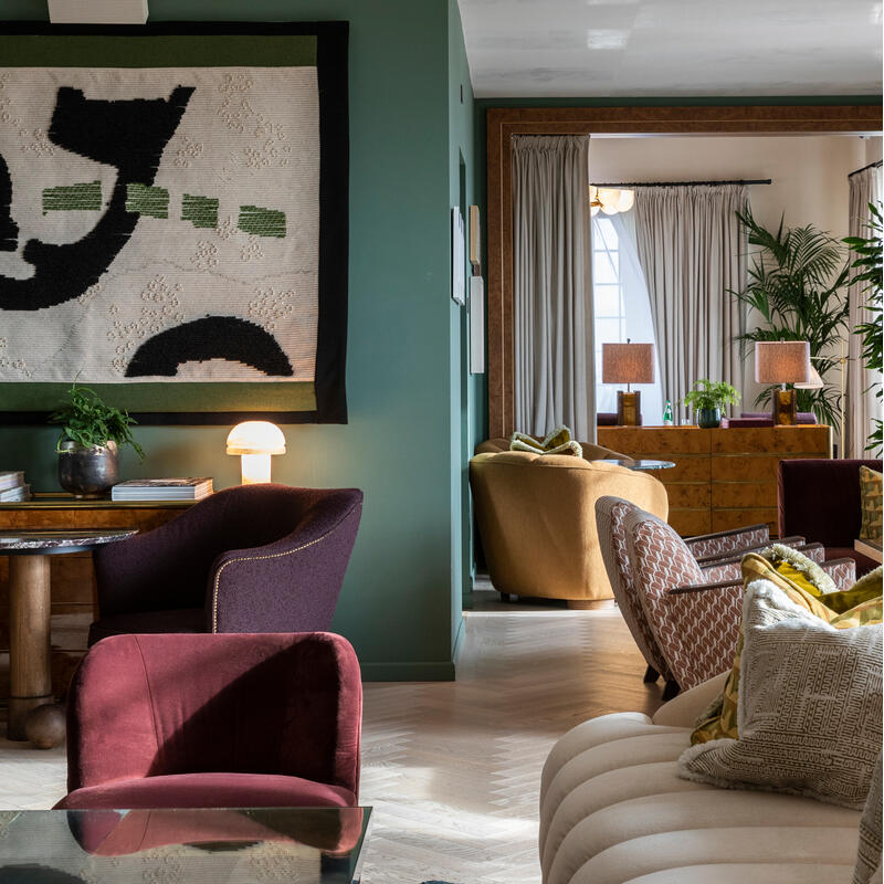 Cozy living room; two purple chairs and table in foreground, modern abstract art on green wall, cushioned sofas and plants in background. Soft lighting, wooden floor, neutral-toned drapery.