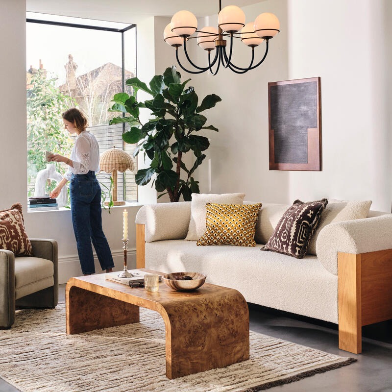 A woman stands by a large window arranging items, surrounded by a cozy living room with a beige sofa, armchair, decorative cushions, wooden coffee table, potted plant, and artwork on the walls.