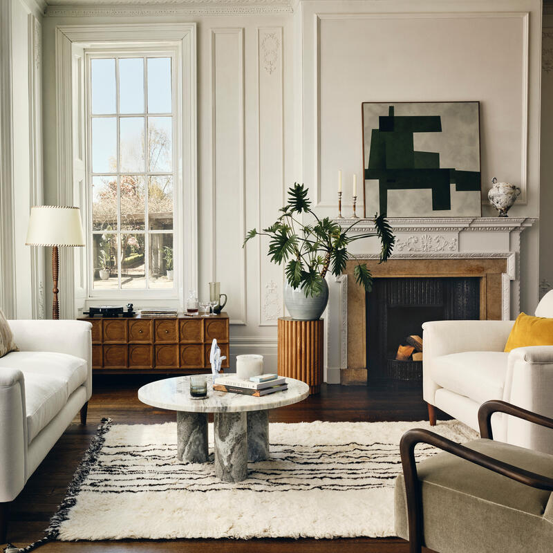 Two white sofas and modern chairs surround a marble coffee table in a bright, elegant living room with large windows, a patterned rug, potted plants, a fireplace, and abstract artwork.