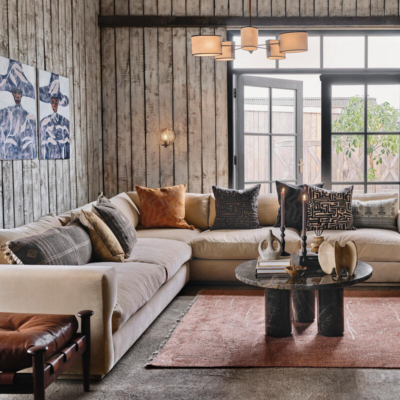 A beige sectional sofa with patterned cushions sits in a cozy, rustic living room featuring wood-paneled walls, a dark round coffee table, a red rug, indoor plants, art, and soft lighting.