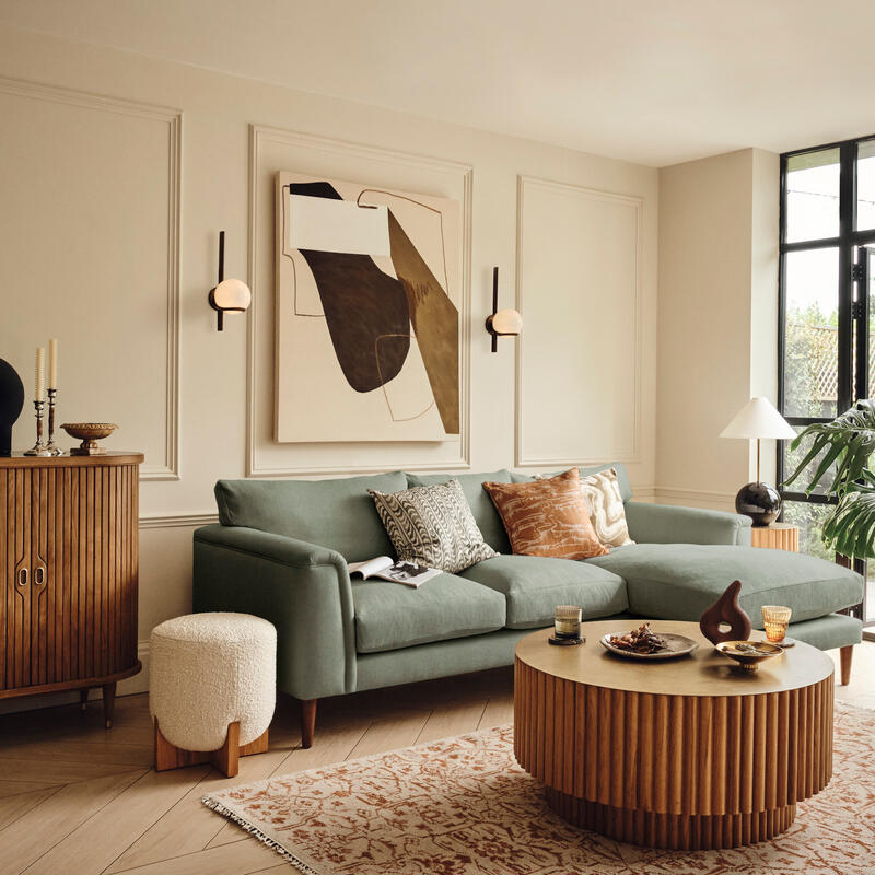 A green sectional sofa with decorative pillows sits in a stylish living room with wooden furniture, abstract wall art, indoor plants, and large windows letting in natural light.