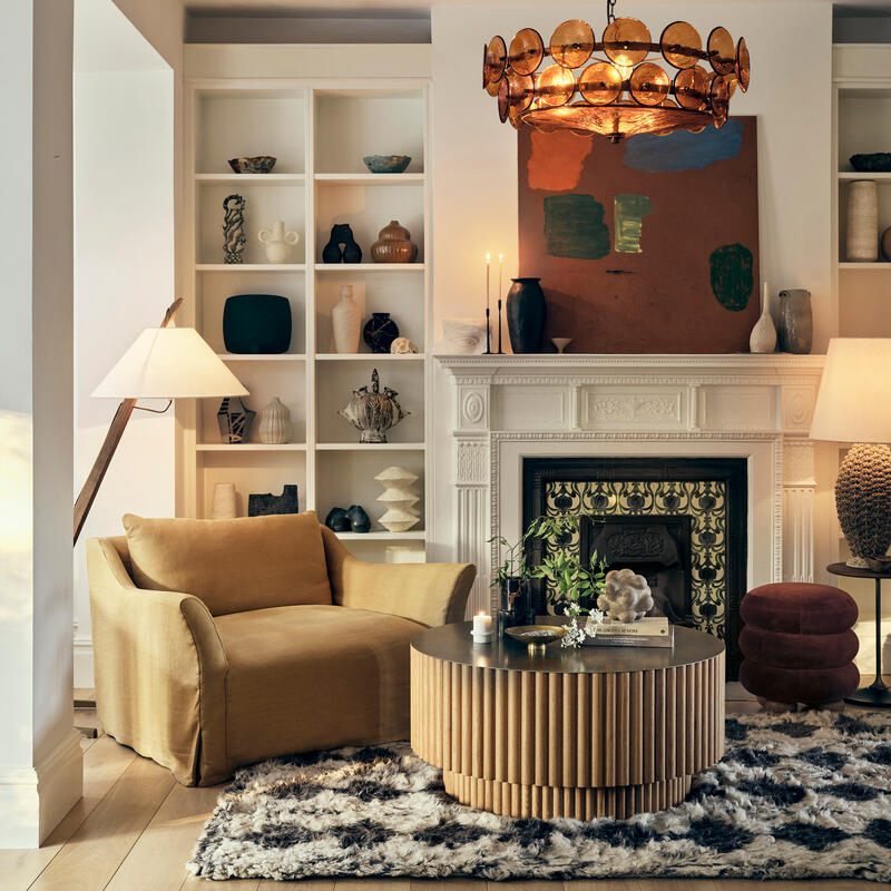 A cozy living room with a mustard-yellow armchair facing a round, wooden coffee table atop a fluffy rug. The room features built-in shelves, a decorative fireplace, and a statement chandelier.