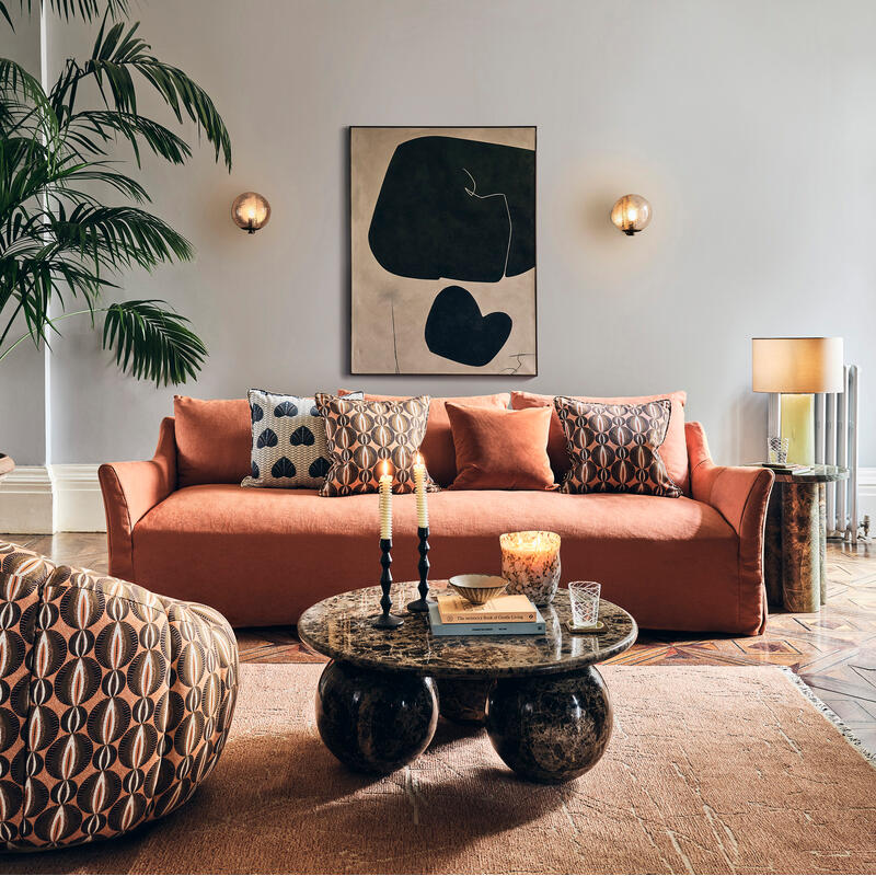 A rust-colored sofa adorned with patterned cushions sits in a spacious room with a large potted plant, abstract wall art, and a marble coffee table holding books and candles.