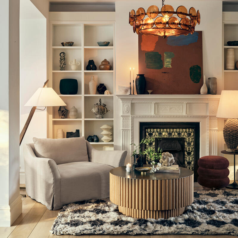 A cozy, modern living room features a beige armchair beside a circular wooden coffee table with decorative items and candles on a fluffy rug, with bookshelves, artwork, and a chandelier in the background.